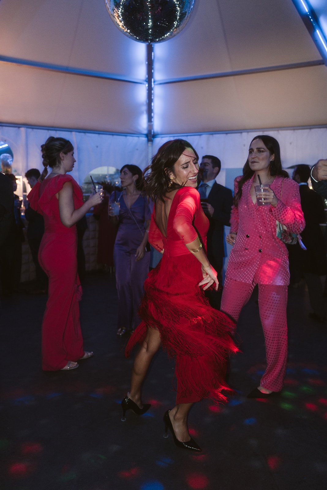 Personas bailando en una fiesta con vestidos elegantes, luces de colores y una bola de discoteca en el techo.