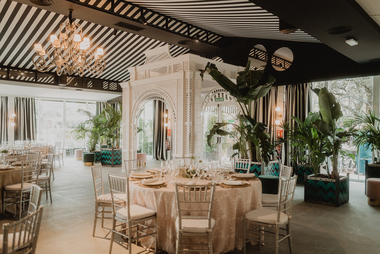 Interior de un elegante salón de eventos con mesas redondas, sillas plateadas, plantas decorativas y una araña colgante. Una estructura blanca detallada se encuentra en el centro, y el techo tiene un diseño de rayas. Hay ventanas grandes y cortinas a