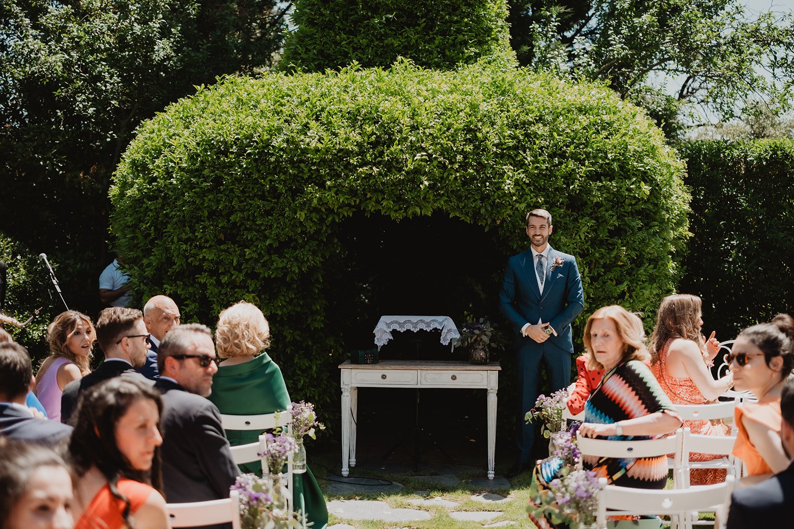 Ceremonia al aire libre con personas sentadas, un hombre de pie con traje azul y un fondo de vegetación.