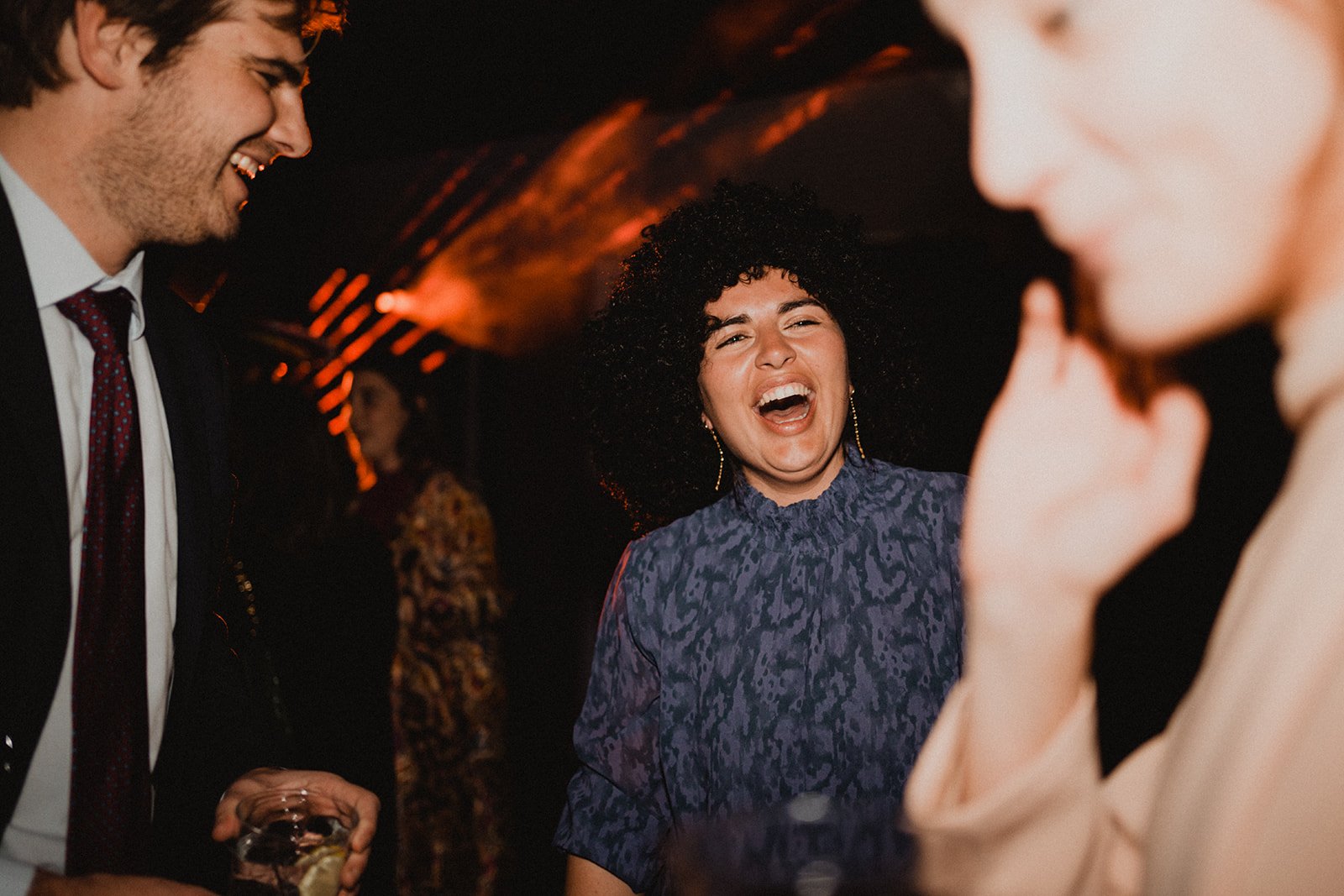 Grupo de personas divirtiéndose en una fiesta, riendo y conversando, con iluminación de fondo. Persona en el centro con cabello rizado sonríe ampliamente.