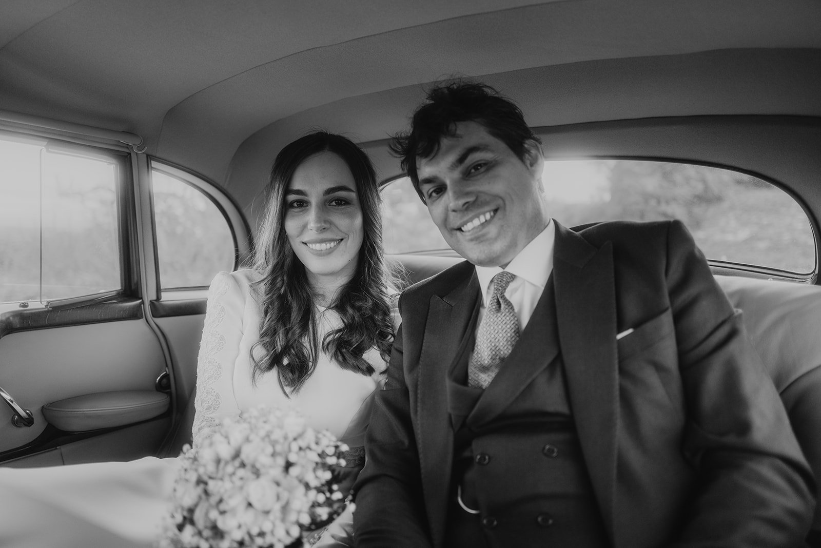 Pareja sonriente en auto, vestidos elegantemente, con un ramo de flores.