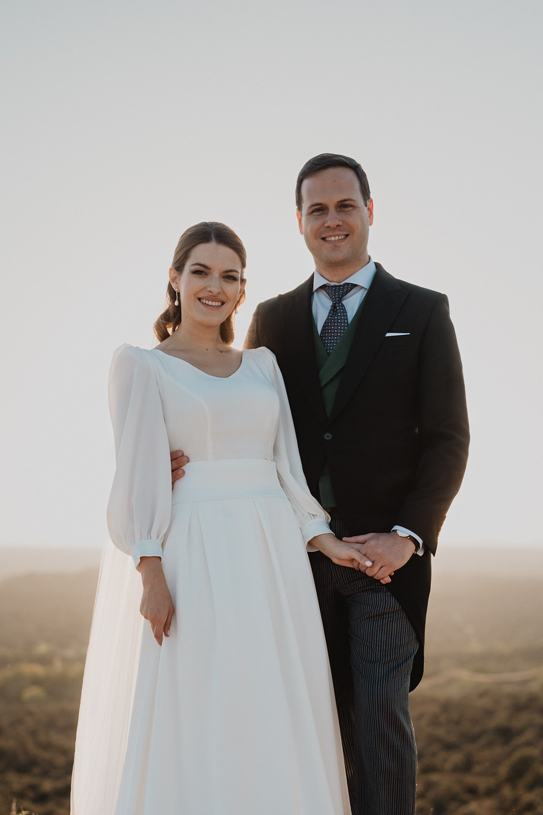 Pareja vestida formalmente, ella con vestido blanco largo y él con traje oscuro, sonríen al aire libre.