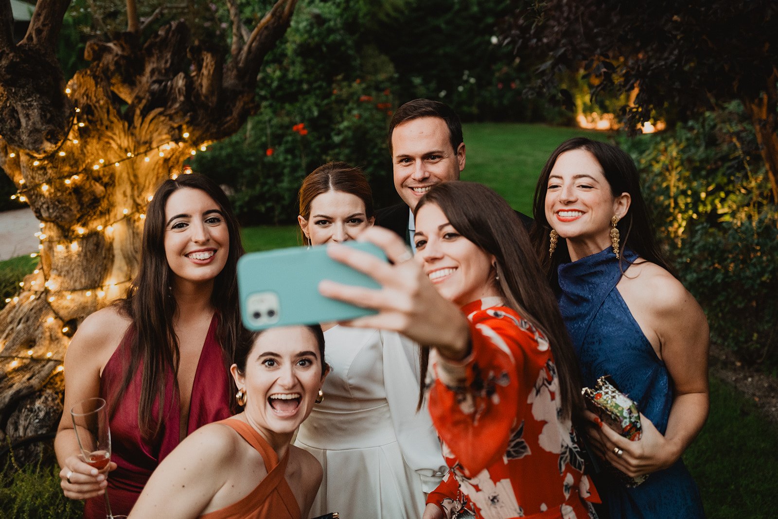 Grupo de personas sonrientes tomando una selfie en un jardín con luces de fondo, durante un evento o celebración por la noche.