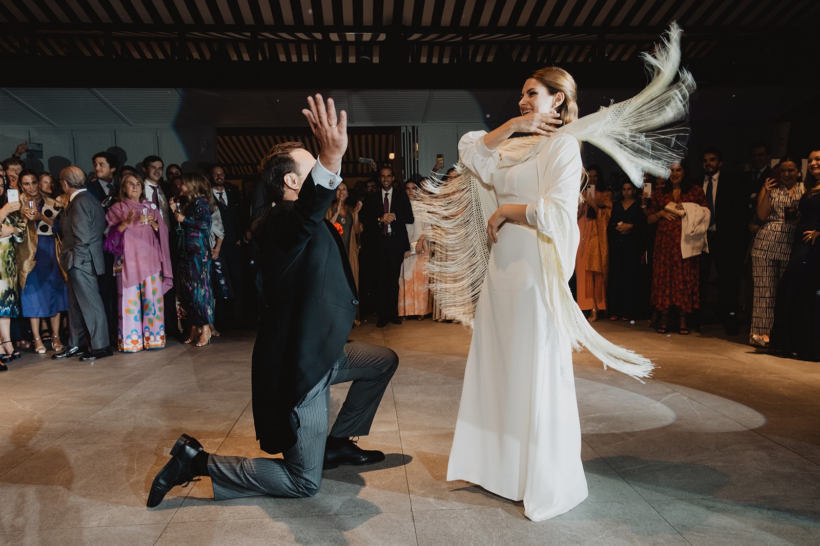 Una pareja bailando en un evento elegante con público observando, el hombre arrodillado y la mujer con vestido blanco y chal.