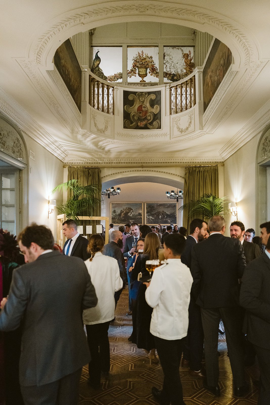 Evento social en un salón elegante con muchas personas conversando y sirviendo bebidas. El techo tiene detalles arquitectónicos decorativos y hay obras de arte en las paredes.