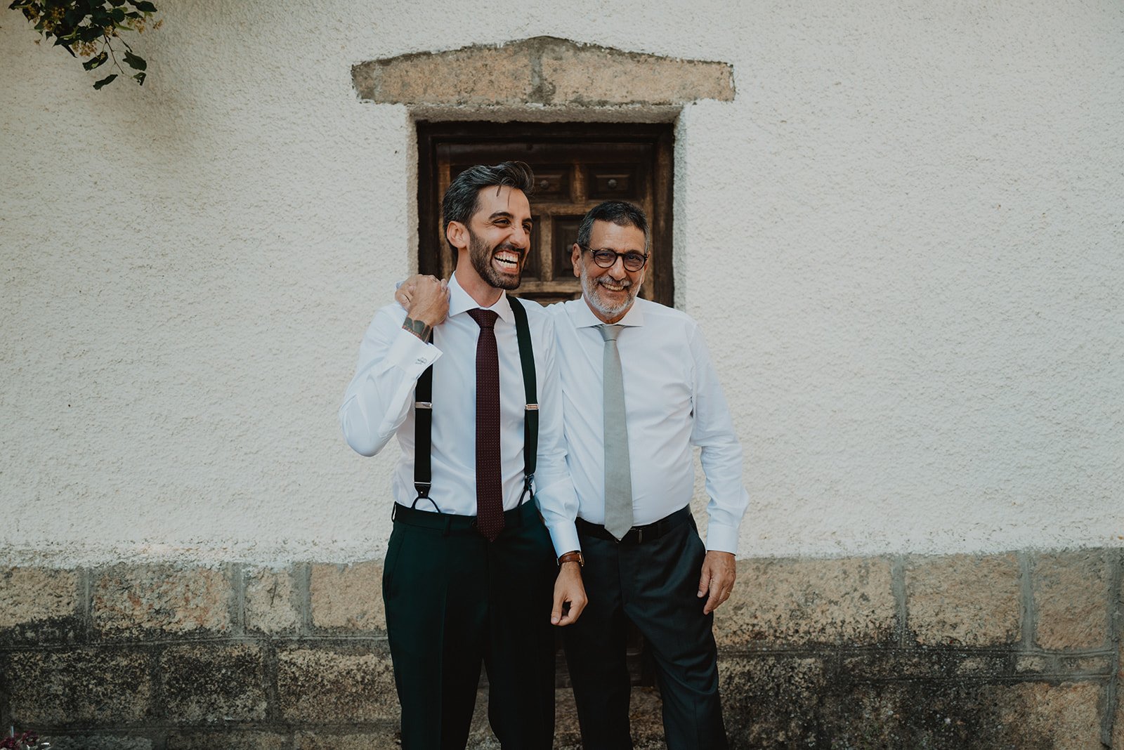 Dos hombres sonrientes con camisas blancas y corbatas junto a una pared blanca y una pequeña ventana de madera.