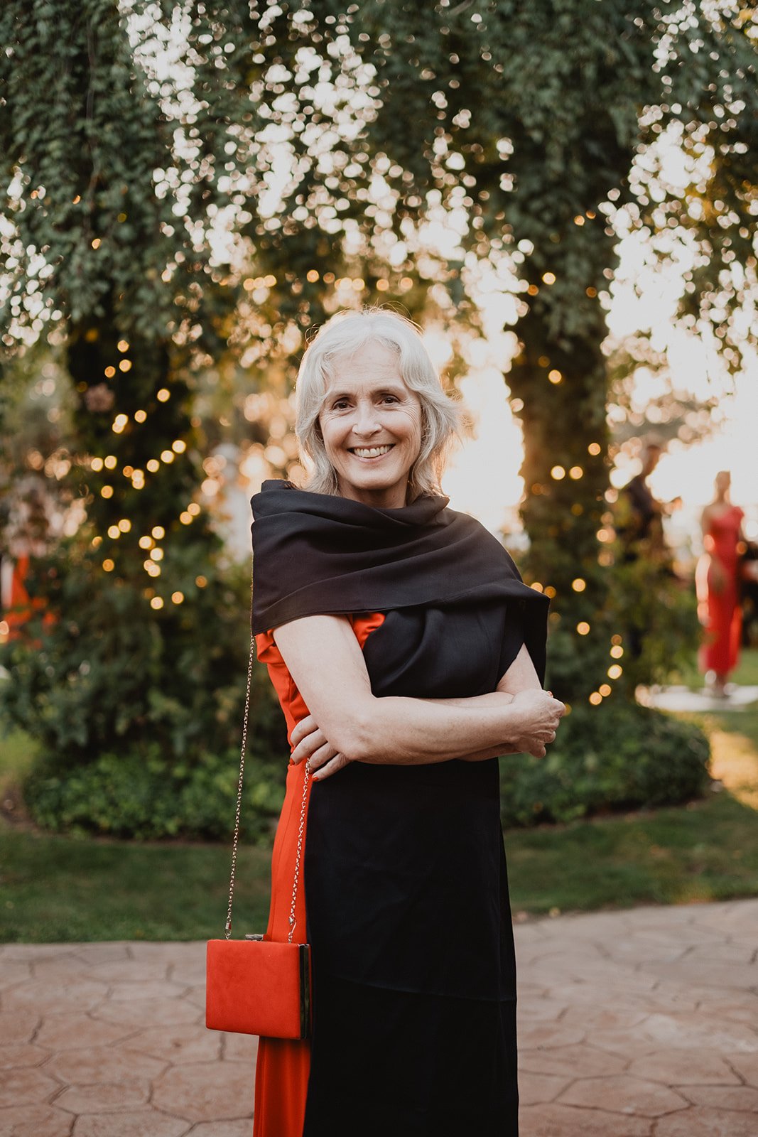 Mujer mayor sonriendo con vestido rojo y chal negro, de pie en un jardín con luces de fondo.