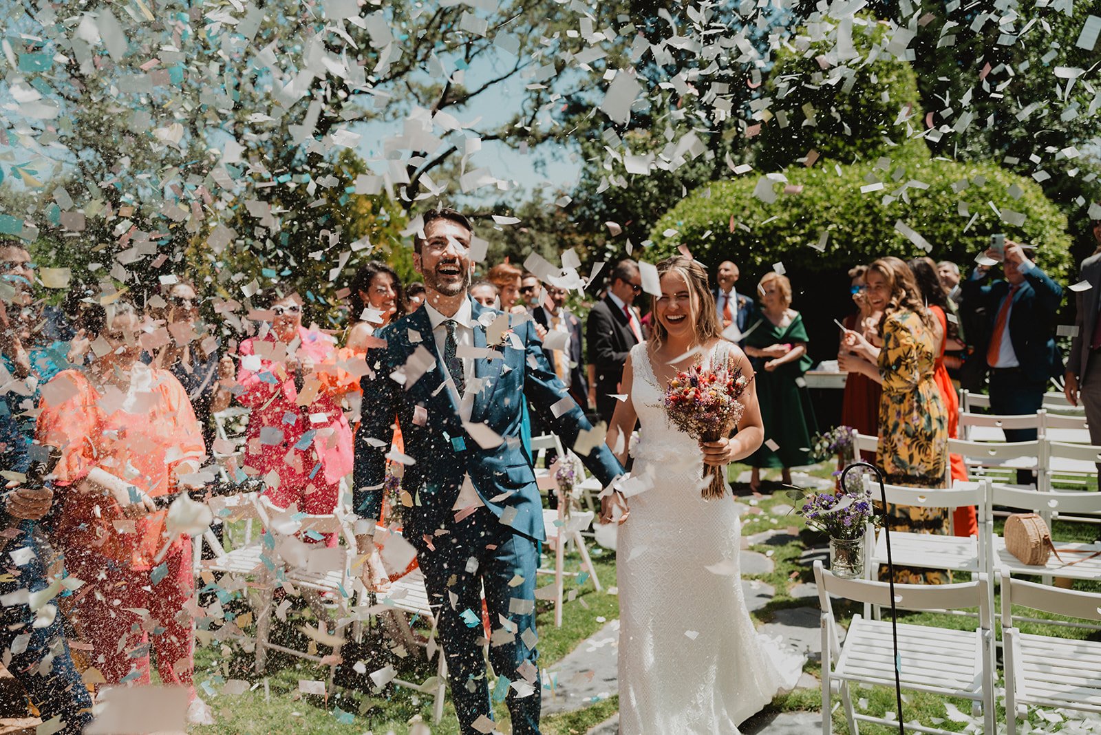 Una pareja recién casada caminando por un pasillo exterior mientras les lanzan confeti, rodeados de invitados felices.