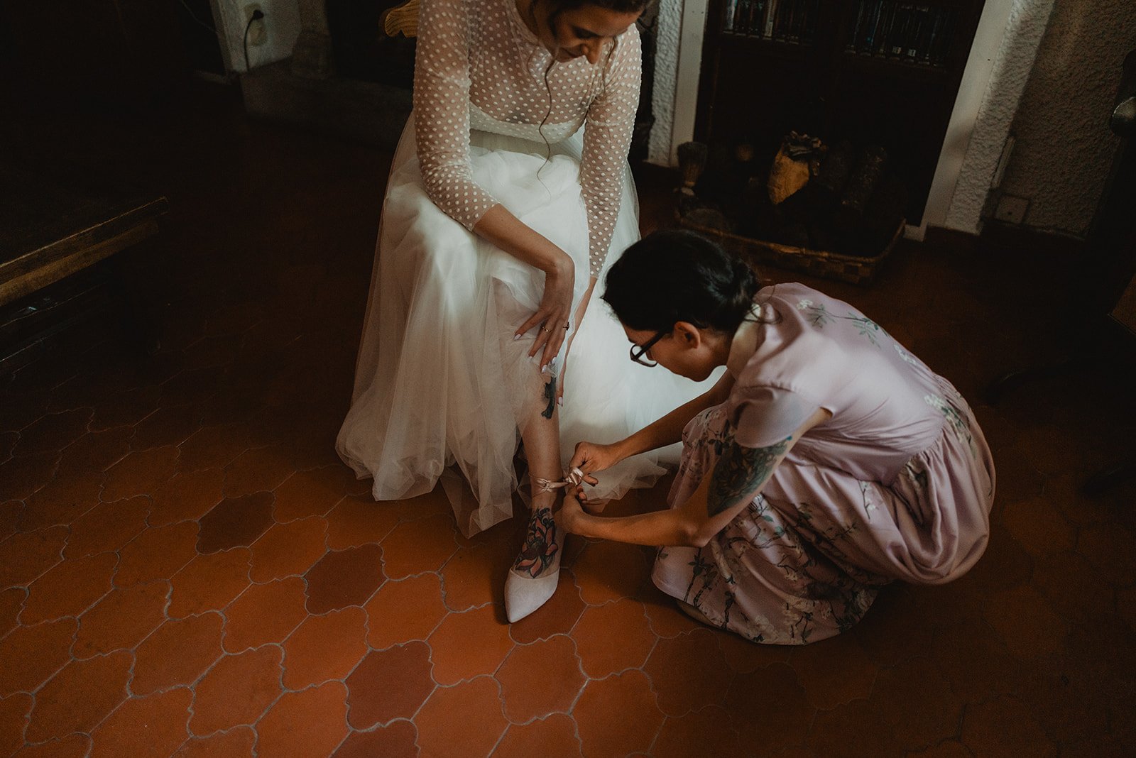 Una novia sentada mientras otra persona le ajusta las cintas de sus zapatos en un ambiente interior con suelo de baldosas.
