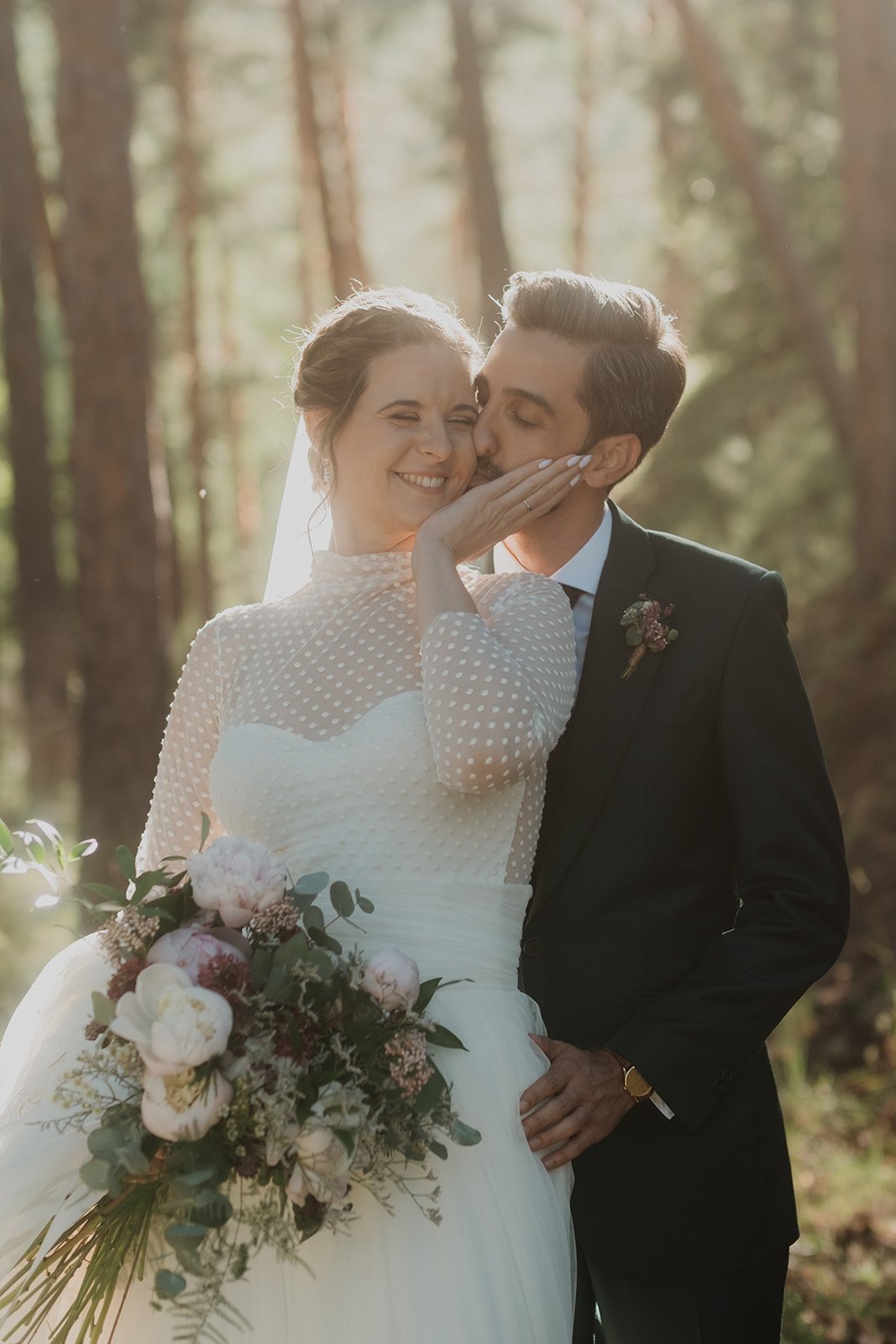 Pareja de novios en el bosque, con el novio besando a la novia en la mejilla y ella sosteniendo un ramo de flores.