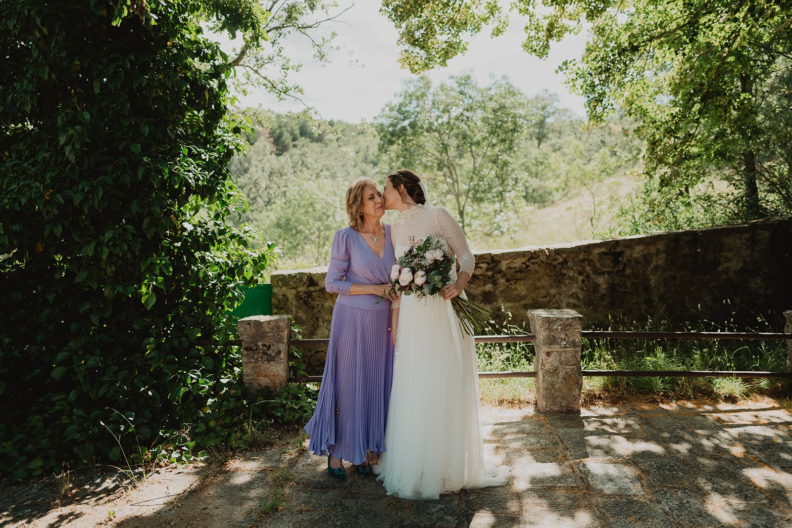 Dos mujeres en un entorno natural, una en vestido de novia blanco besando a la otra que lleva un vestido lila, ambas sonríen.