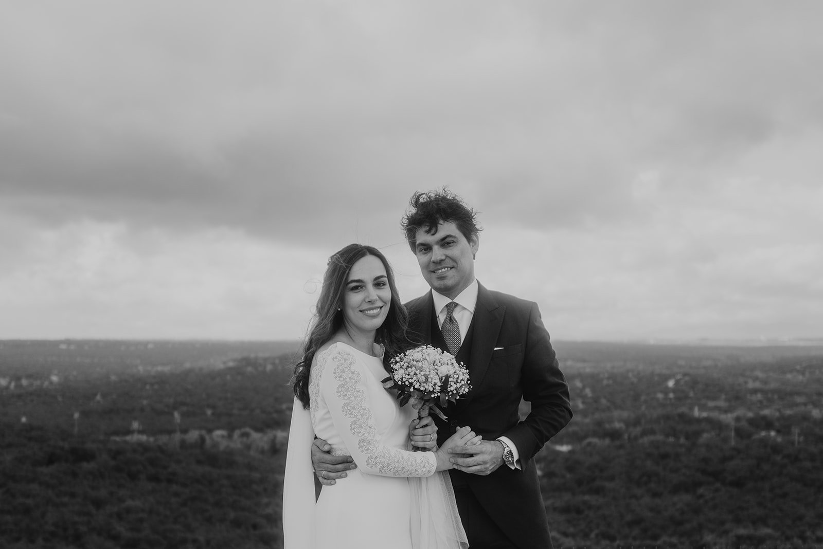 Pareja de novios posando al aire libre, con el novio en traje y la novia en vestido blanco, sosteniendo un ramo de flores.