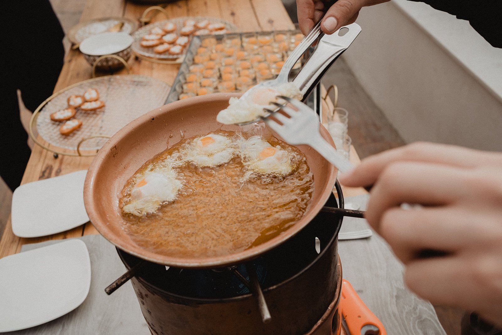 Cocinando huevos fritos en sartén sobre una estufa de gas al aire libre, con platos y bocadillos en la mesa de madera.