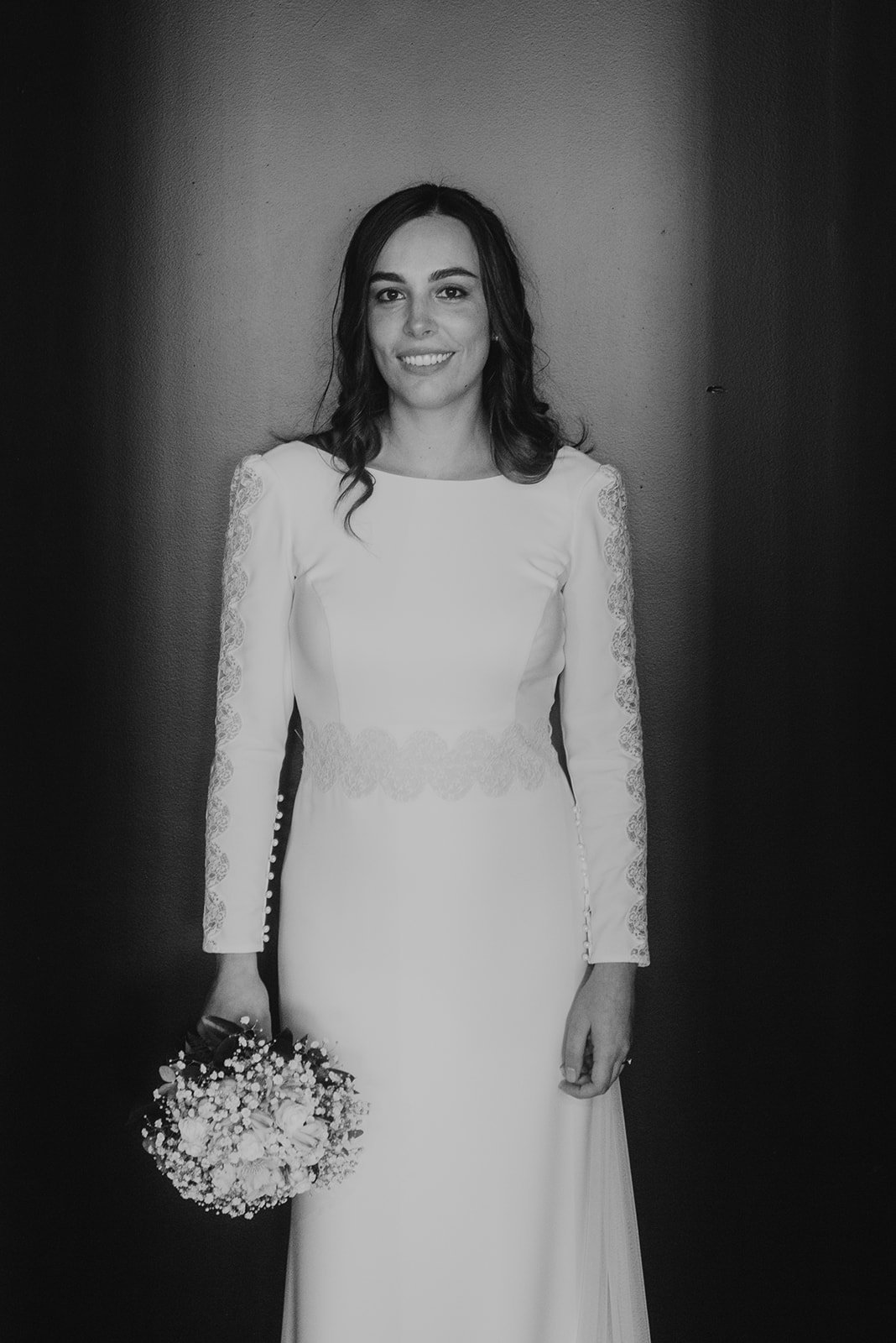 Novia con vestido blanco de encaje, sosteniendo un ramo de flores, fondo oscuro.