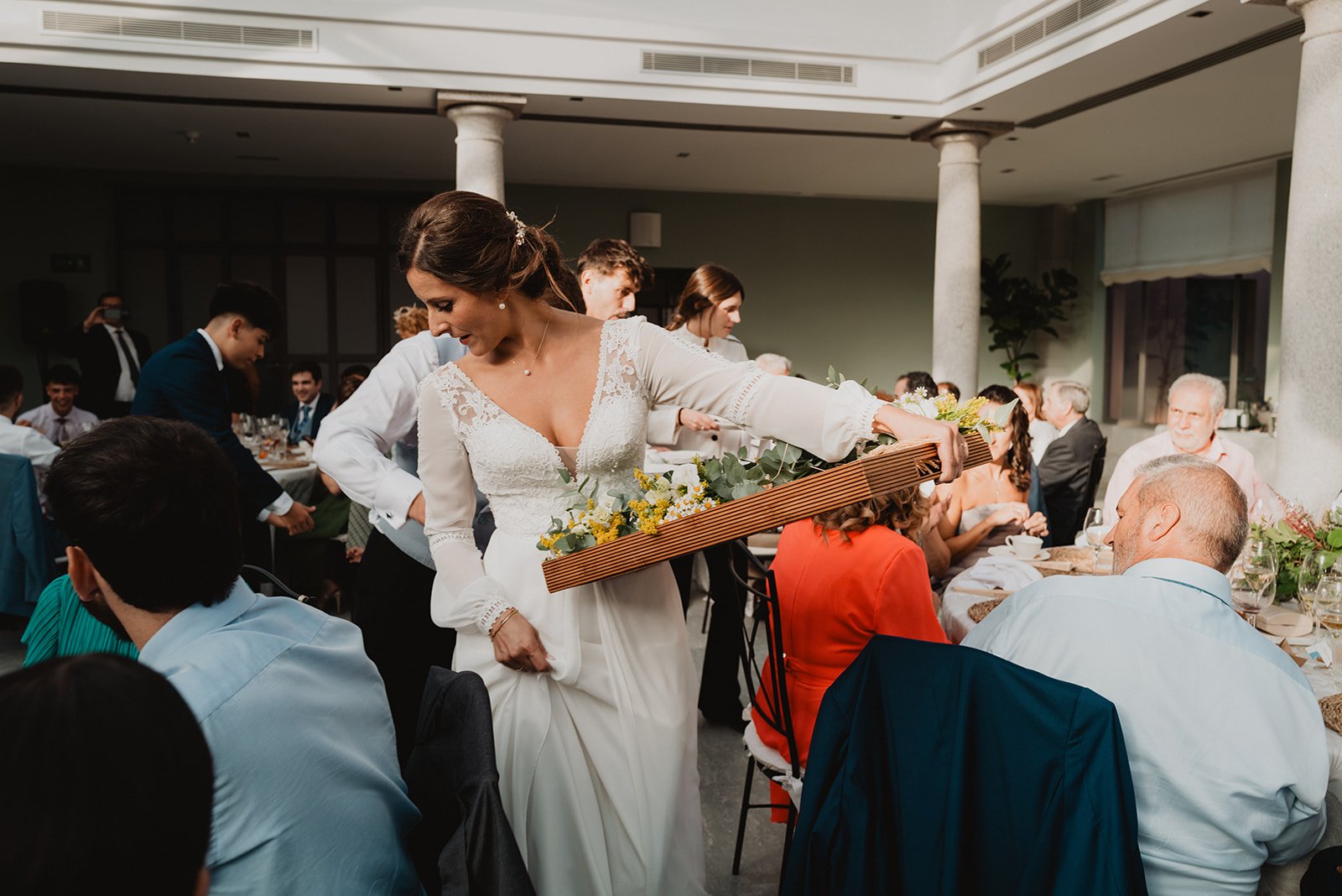Novia en una recepción de boda entregando regalos, rodeada de invitados sentados en mesas con decoración floral.