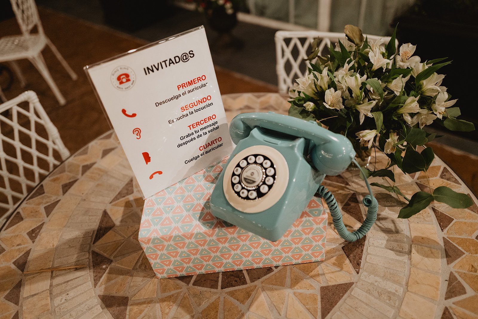 Teléfono vintage azul en una mesa junto a un ramo de flores y un letrero con instrucciones en español para invitados.