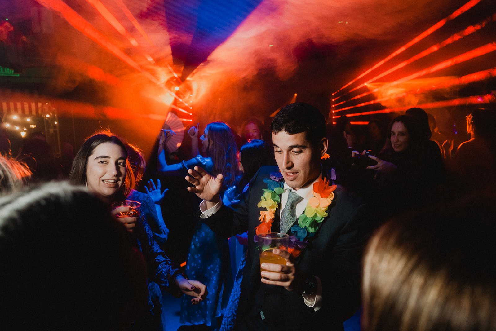 Personas bailando en una fiesta con luces de colores y humo, uno de ellos con un collar de flores y un vaso en la mano.
