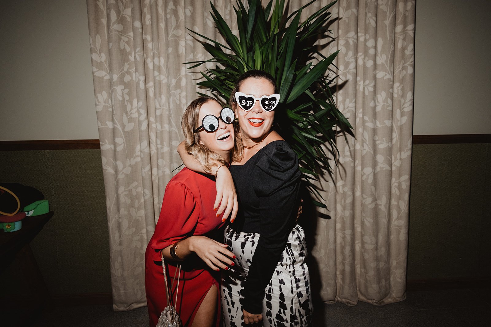 Dos mujeres sonrientes abrazándose, usando disfraces graciosos de gafas grandes frente a una cortina decorativa.