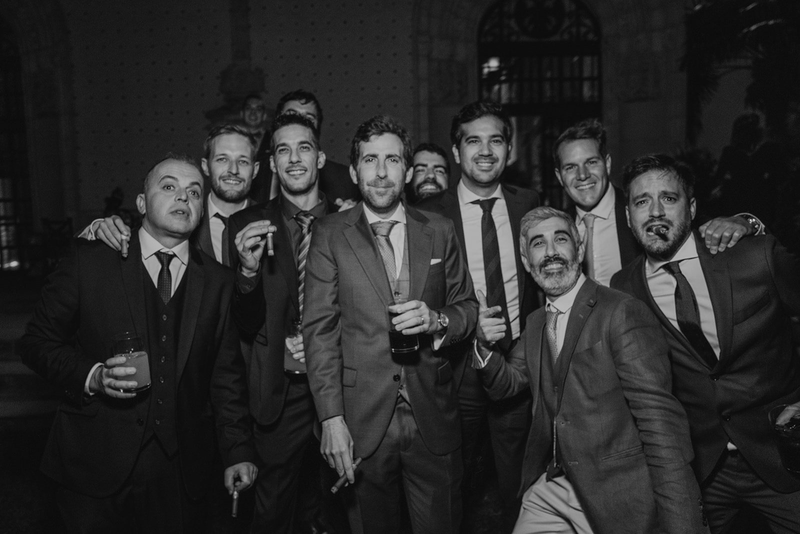 Grupo de hombres en trajes posando y sonriendo, algunos sostienen puros y bebidas, en un ambiente de fiesta nocturna.