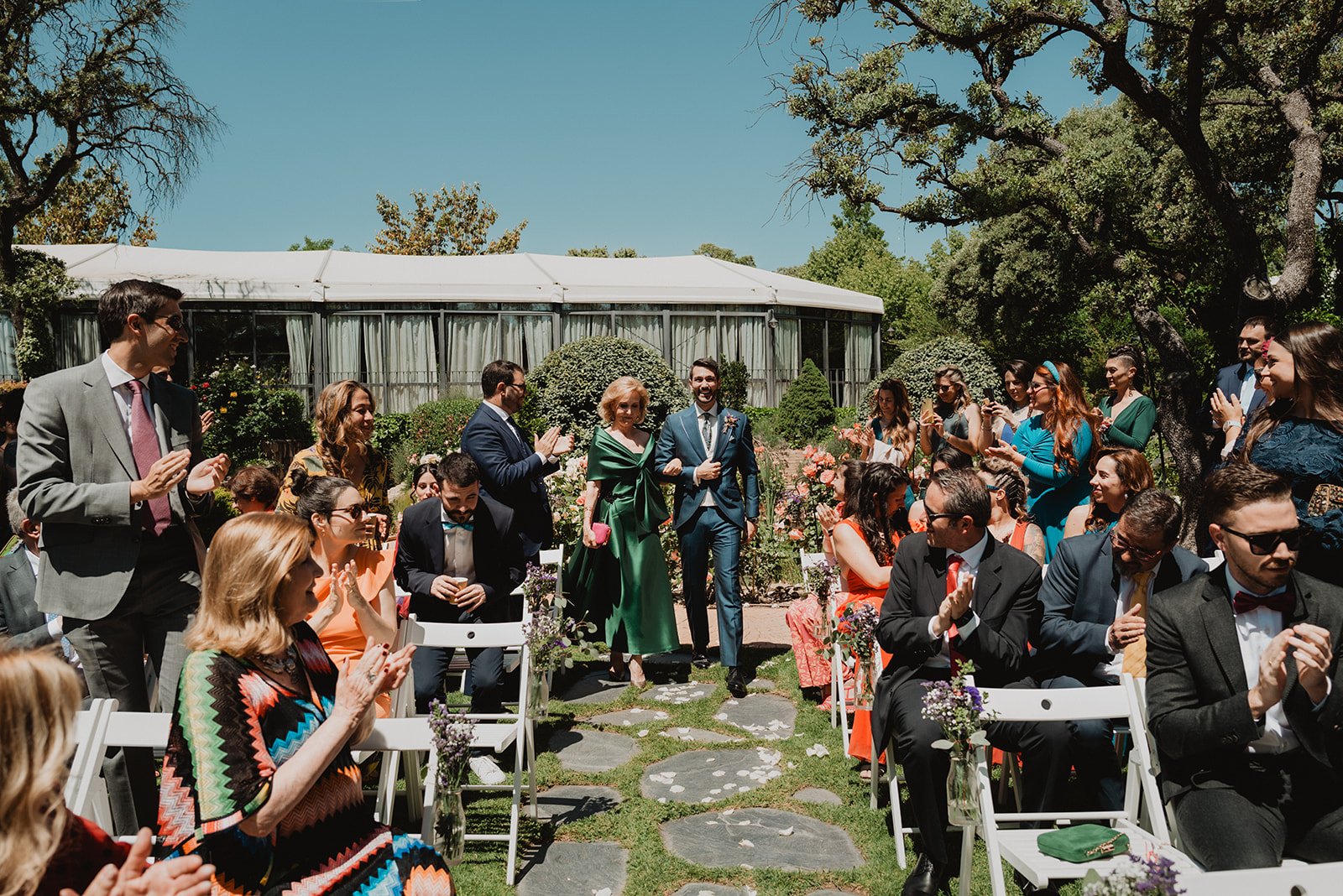 Ceremonia de boda al aire libre con personas sentadas en sillas, aplaudiendo mientras una pareja camina por el pasillo central. El entorno tiene árboles y flores, y hay un edificio detrás.