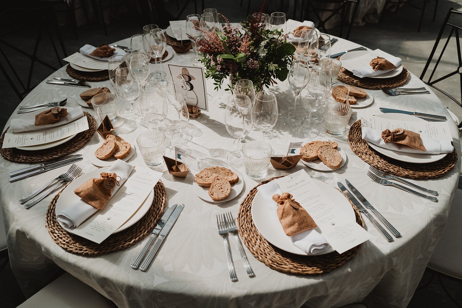 Mesa redonda elegantemente decorada para un evento, con platos, cubiertos, copas, servilletas, pan y centros de mesa florales.