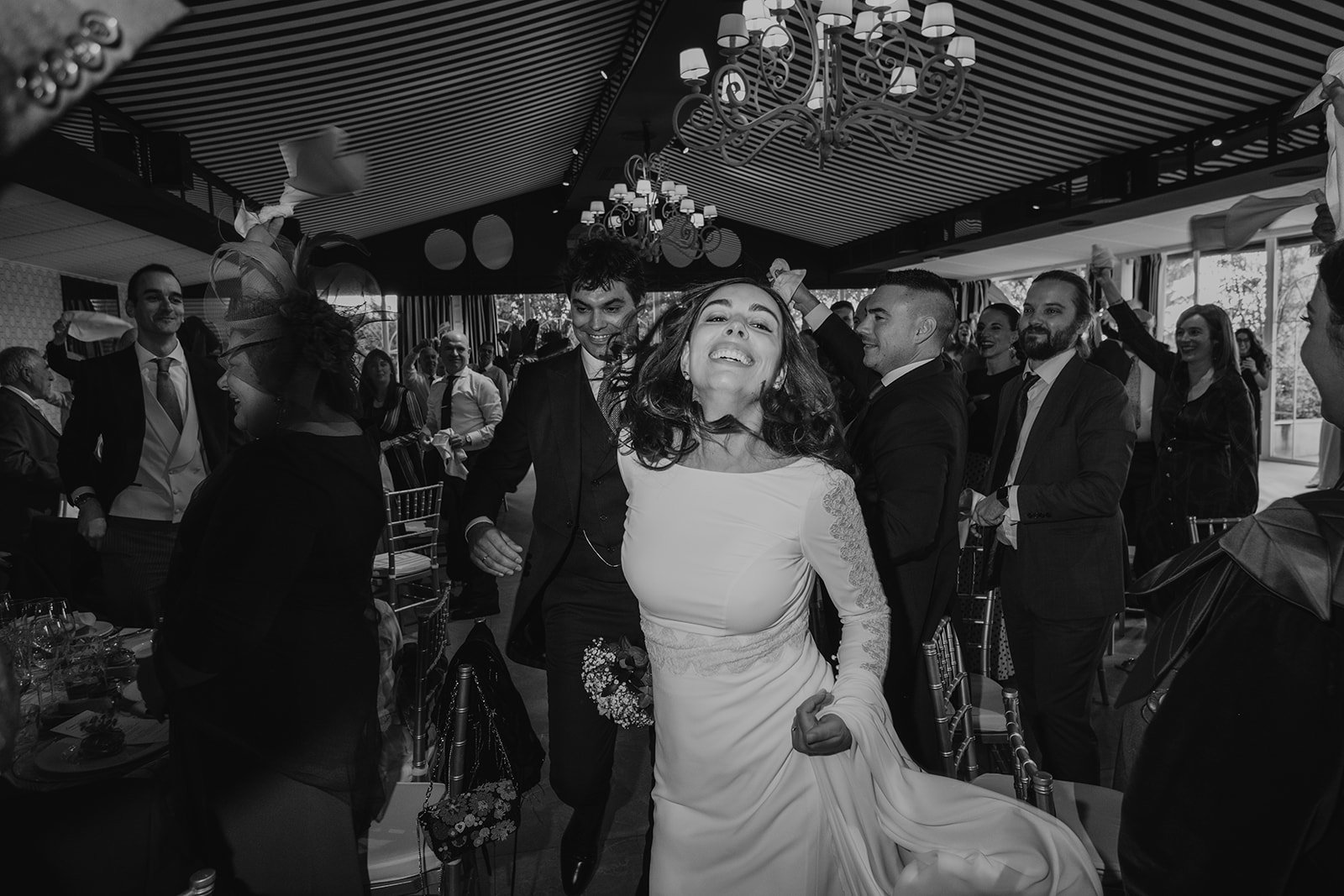 Pareja de recién casados bailando felices en una recepción de boda, rodeados de invitados que celebran con pañuelos en el aire. Ambiente festivo en blanco y negro con decoración elegante.