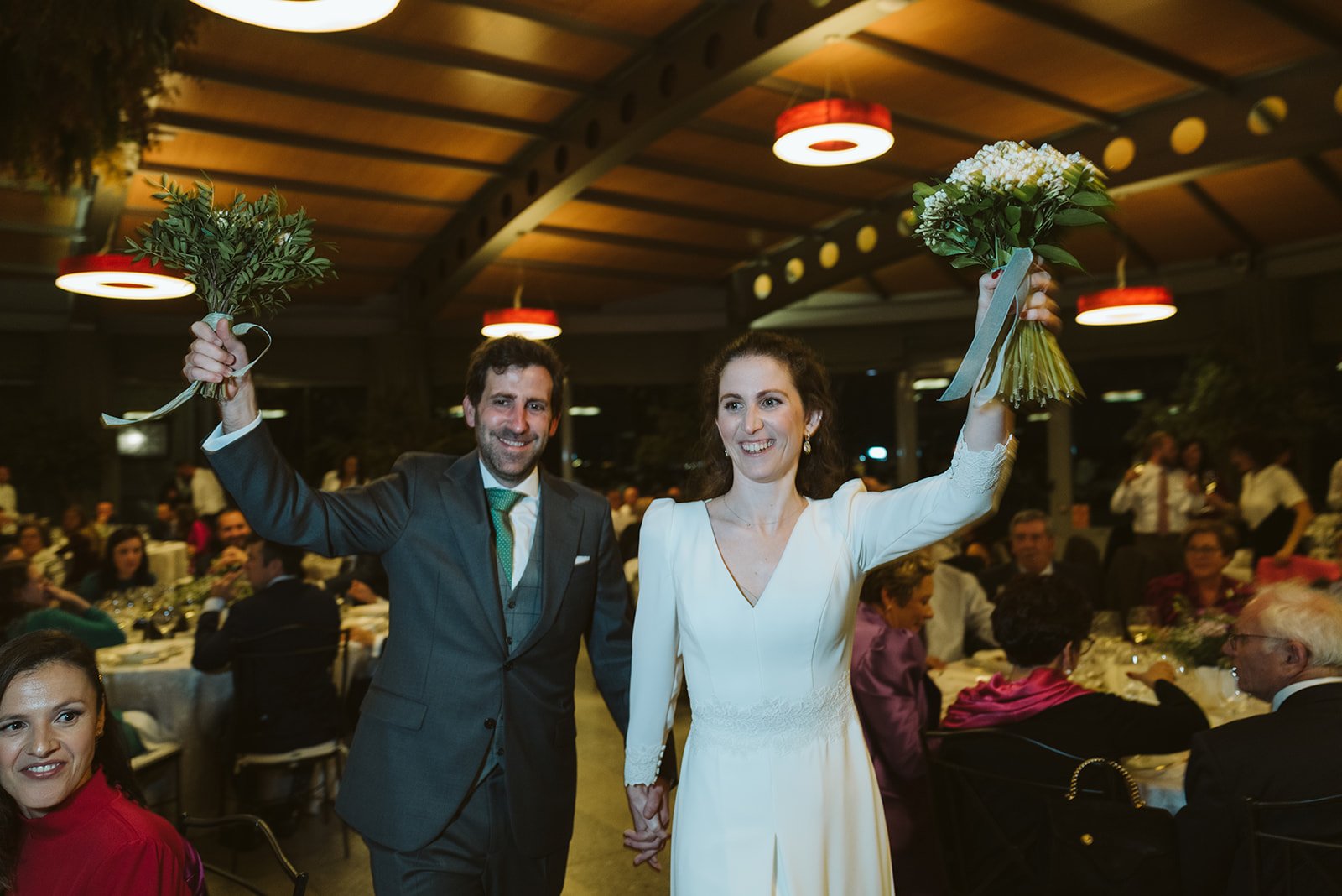 Una pareja sonriente vestida de traje y vestido de novia, con ramos de flores en las manos, celebra en una recepción de boda rodeada de invitados sentados en mesas.