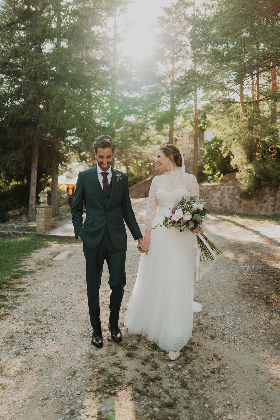 Pareja de novios caminando de la mano en un camino rodeado de árboles, ella con vestido de novia y ramo de flores, él con traje oscuro. Fondo de bosque iluminado por el sol.