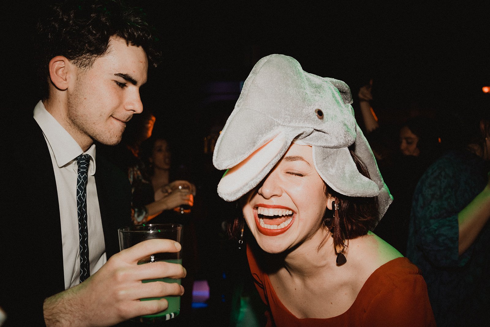 Dos personas en una fiesta, una mujer con sombrero de elefante, ambos sosteniendo bebidas y riendo.