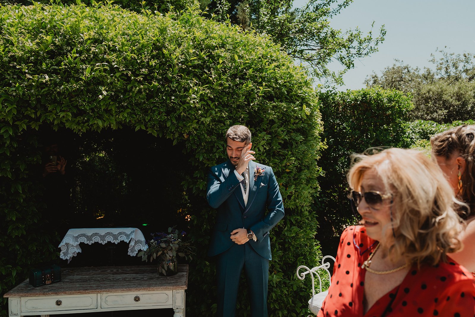 Persona en traje azul en un jardín, rodeado de arbustos verdes. Mesa decorada con mantel blanco y flores. Mujer de cabello rubio con gafas de sol en primer plano.