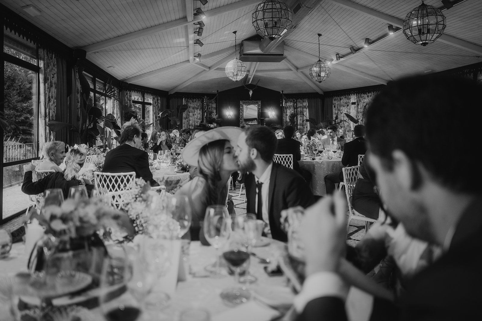 Pareja besándose en una recepción de boda, con invitados sentados en mesas elegantemente decoradas, en un salón amplio y bien iluminado.