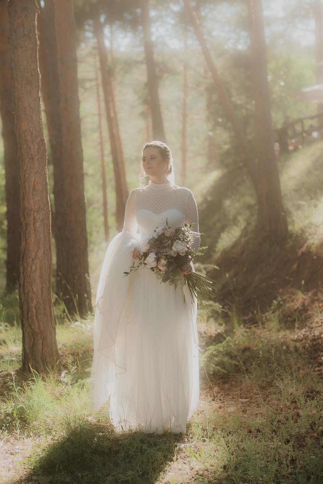 Novia en vestido blanco sosteniendo un ramo de flores en el bosque, iluminada por luz solar.