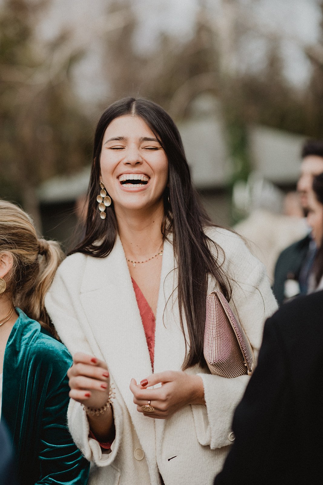 Persona sonriendo en una reunión social, con un abrigo blanco, pendientes grandes y un bolso de mano. Fondo con más personas y un ambiente al aire libre.