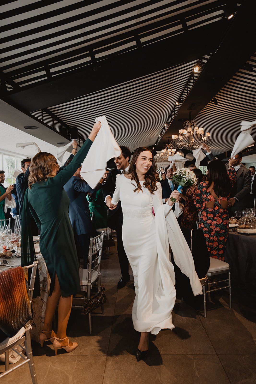 Una novia caminando entre invitados en una recepción de boda, con personas levantando servilletas blancas y aplaudiendo.