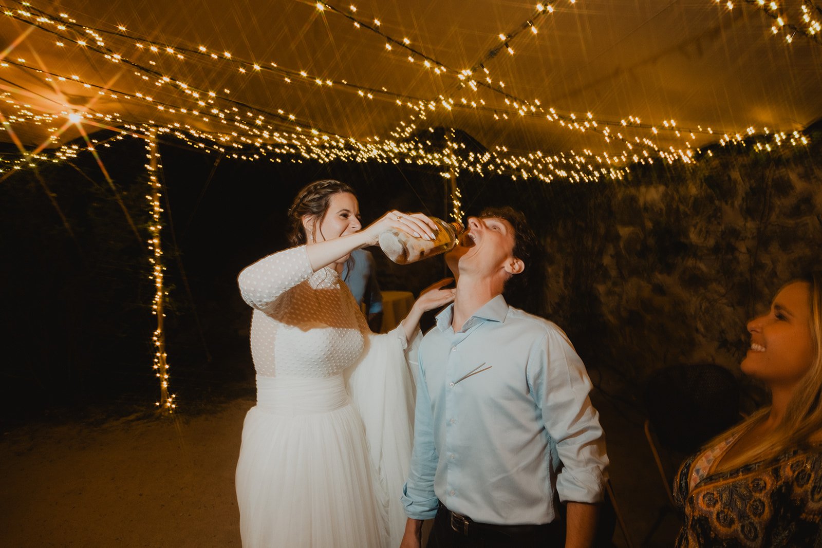 Una persona vestida de novia sirve licor de una botella a otra persona bajo luces decorativas en una fiesta.