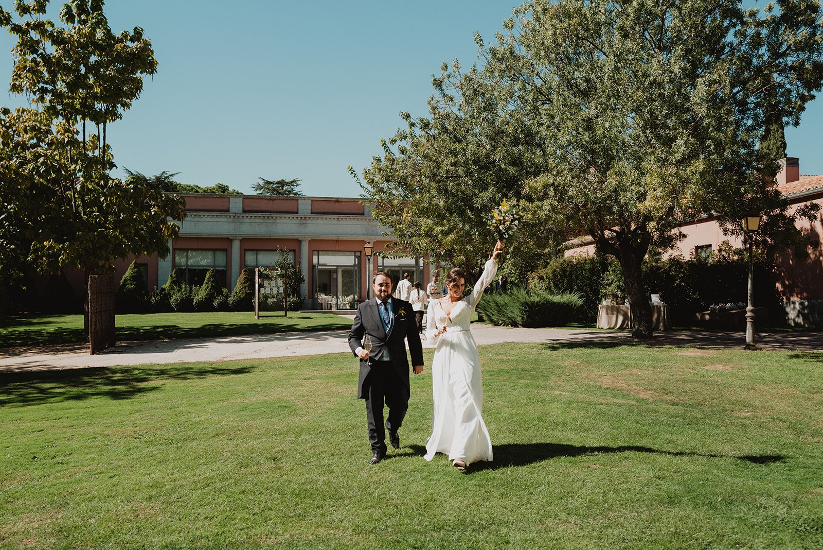 Pareja vestida elegantemente caminando en un jardín, la mujer con un ramo de flores levantado, frente a un edificio y árboles.