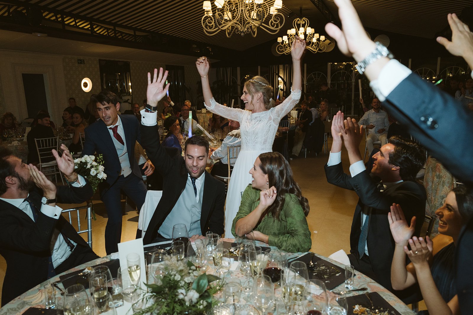 Una celebración de boda en un salón elegante, con varias personas aplaudiendo y levantando las manos alrededor de una mesa. La novia está de pie, sonriendo y levantando los brazos, mientras los invitados muestran entusiasmo y alegría.