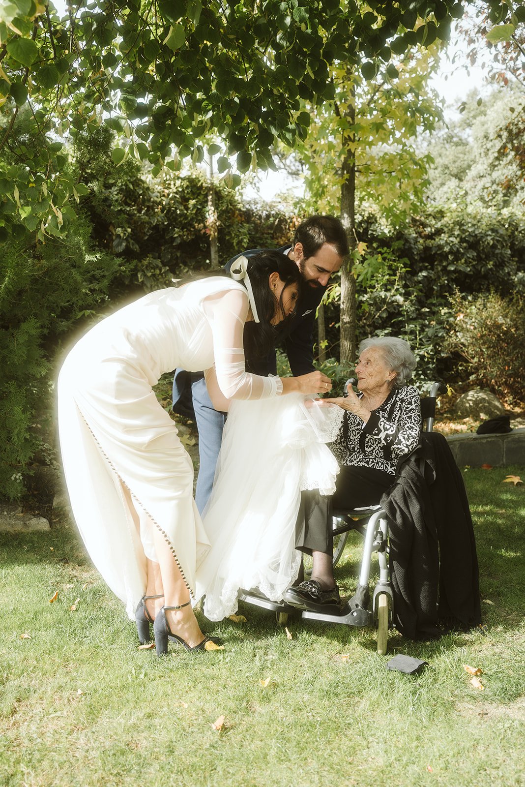 Pareja sonriente en traje de boda junto a una mujer mayor en silla de ruedas, en un jardín.