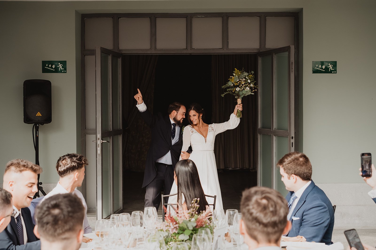 Pareja de recién casados entrando a una recepción con invitados en una mesa, la novia lleva un ramo de flores.