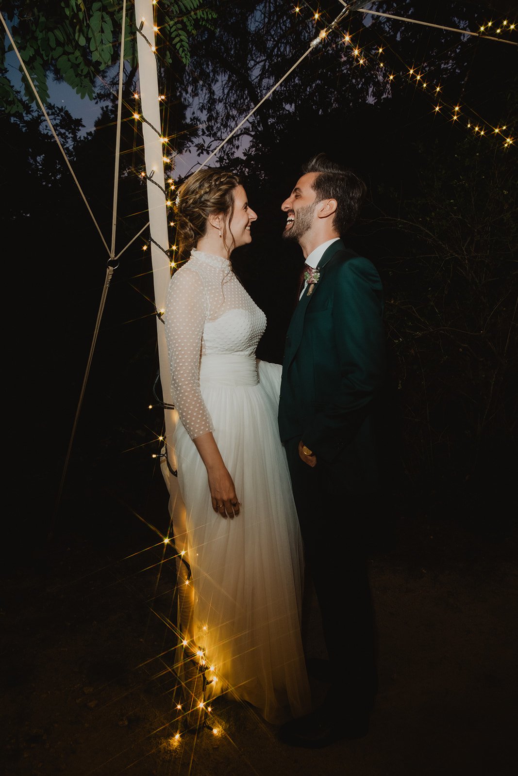 Novia y novio sonriendo de pie bajo luces decorativas en una boda nocturna, rodeados de un ambiente oscuro con árboles.