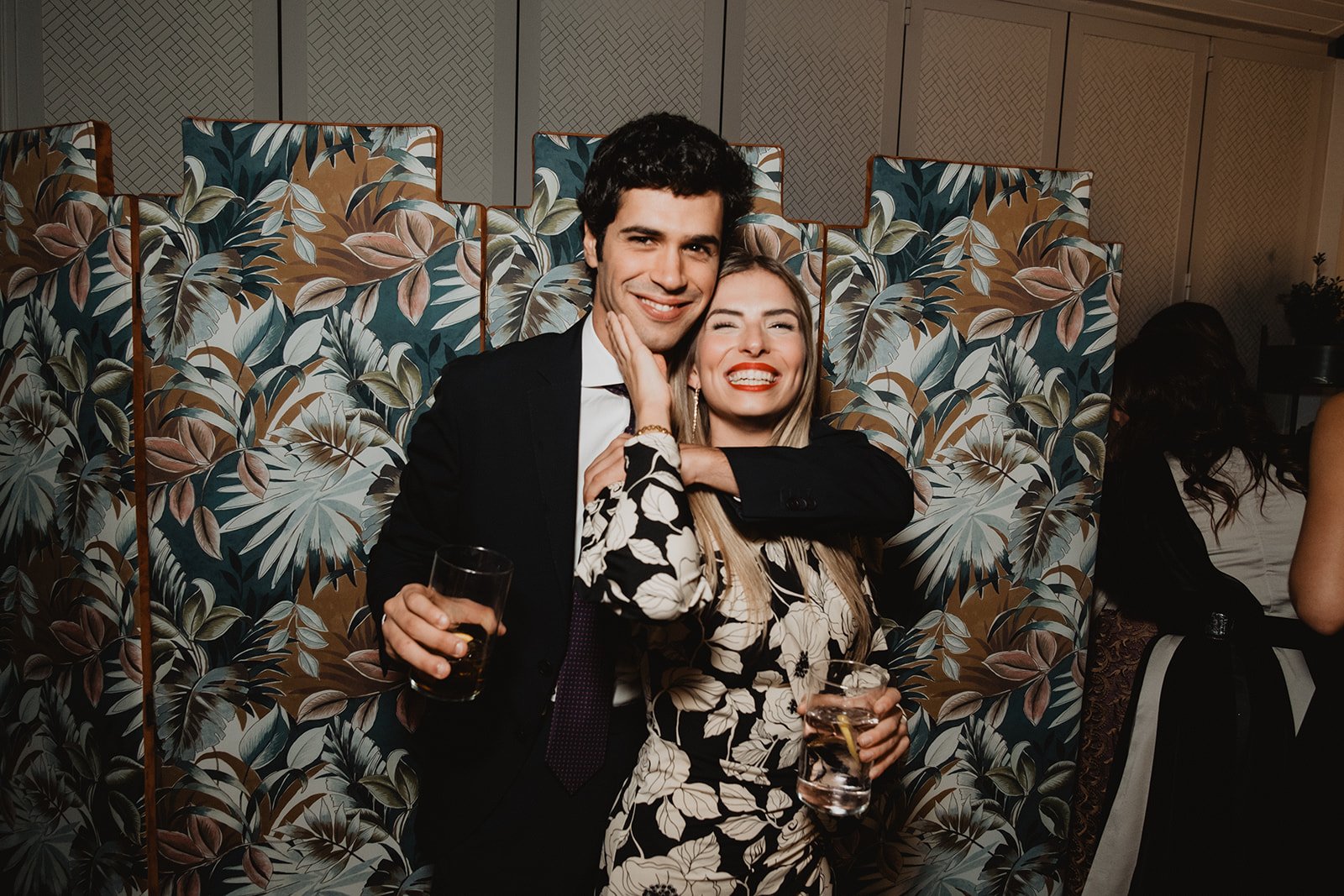 Pareja sonriendo y abrazándose, sosteniendo bebidas, frente a un fondo floral en un evento social.