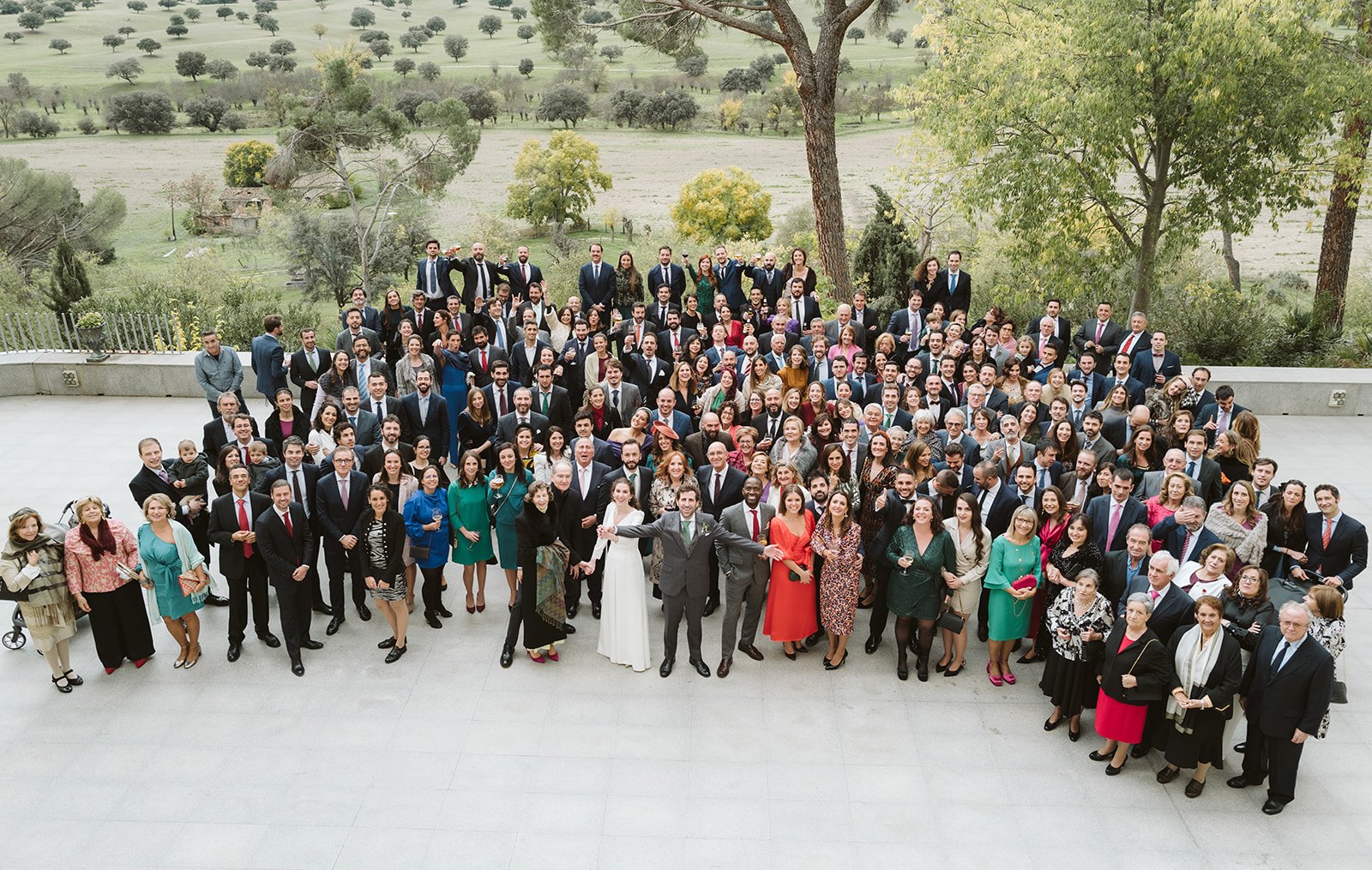 Un grupo grande de personas elegantemente vestidas reunidas al aire libre, posiblemente para un evento especial, con un paisaje verde de fondo.