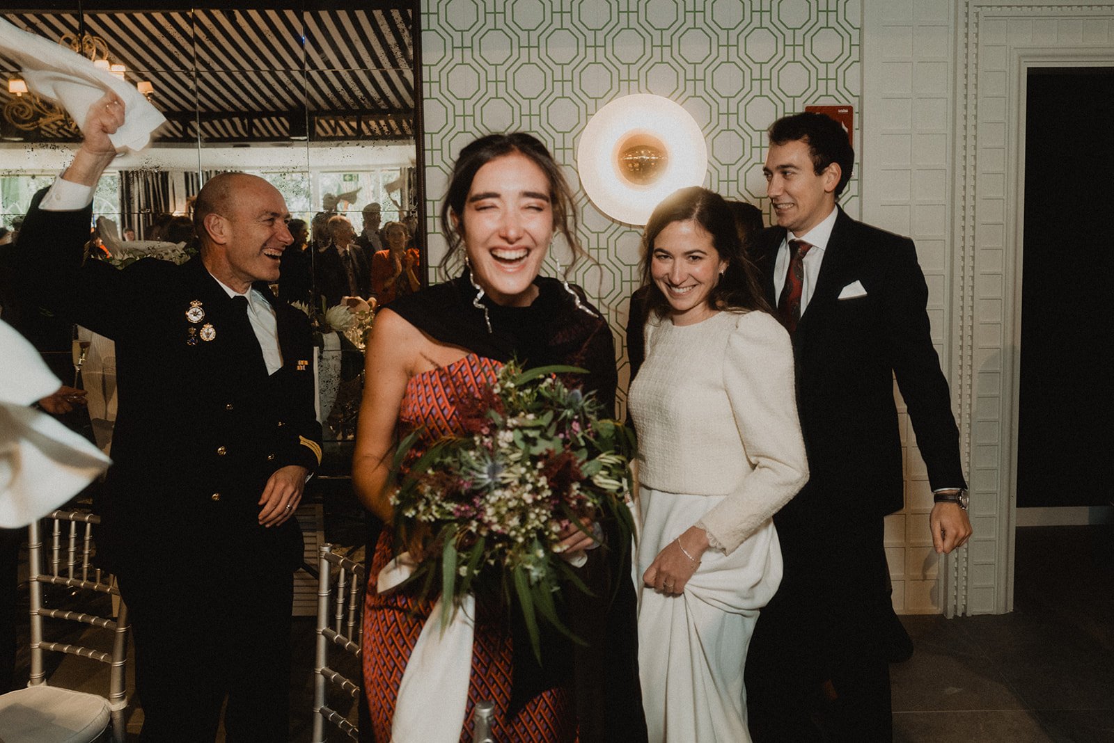 Personas felices en una celebración, una mujer sostiene un ramo de flores, otra mujer a su lado lleva vestido claro, y un hombre con uniforme levanta una servilleta. Decoración elegante y espejos al fondo.