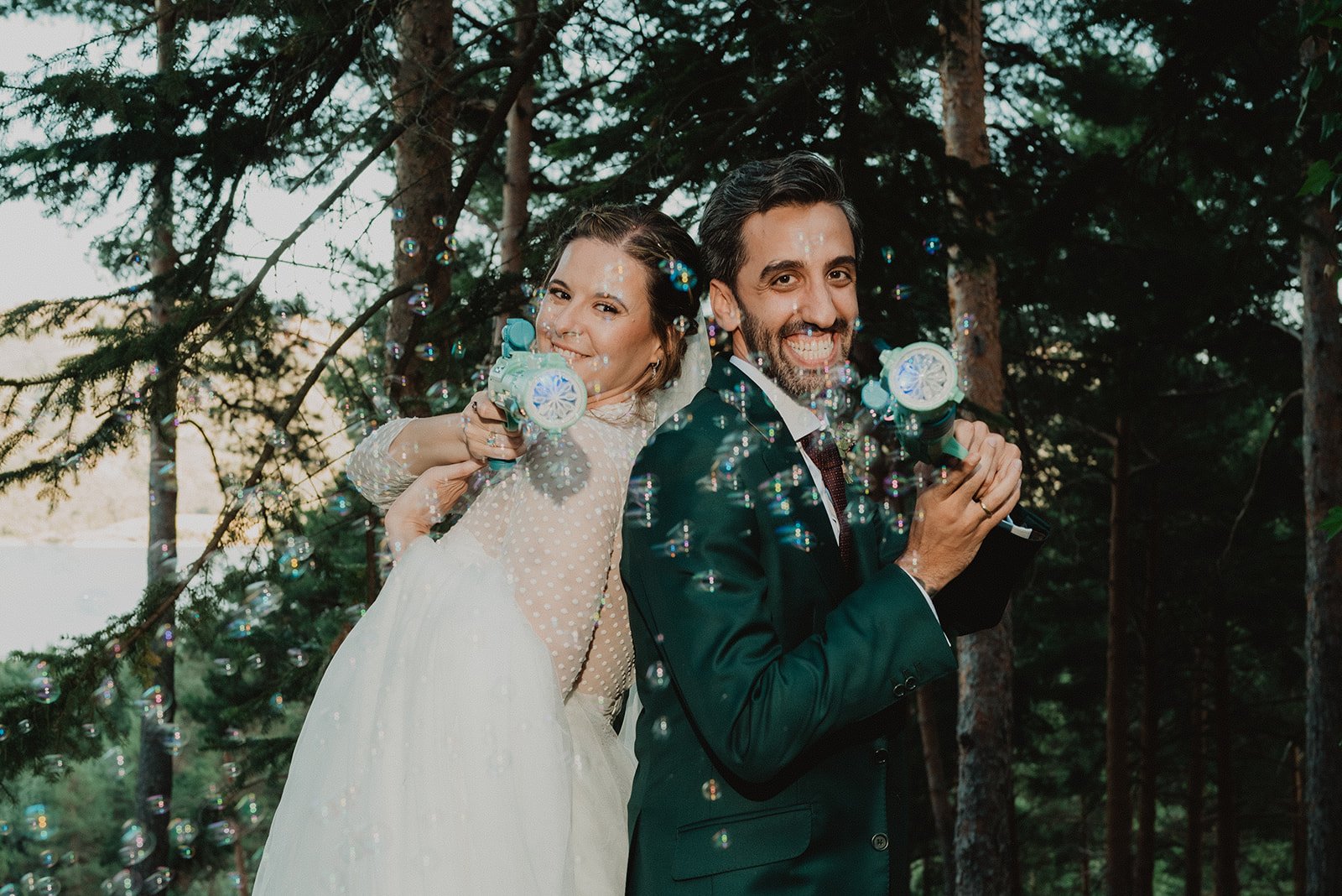 Pareja recién casada en un bosque, sonriendo y usando pistolas de burbujas.