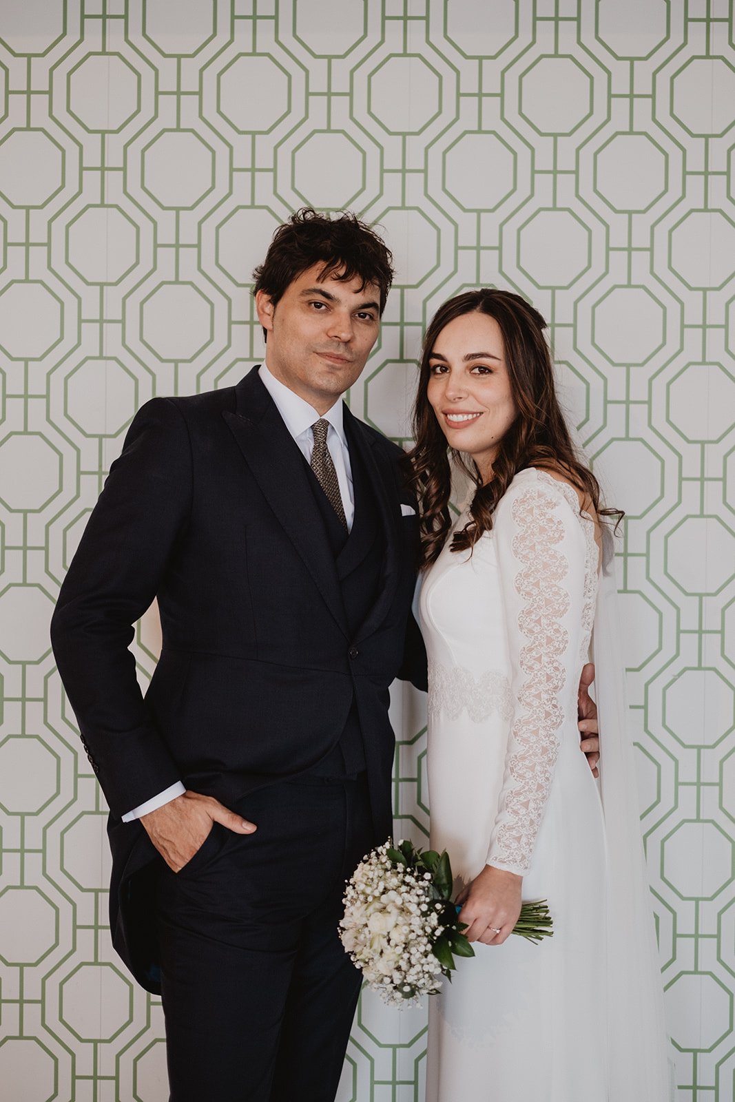 Pareja posando en una boda con fondo geométrico verde. Él lleva traje oscuro, ella lleva vestido de novia blanco con encaje y sostiene un ramo de flores.