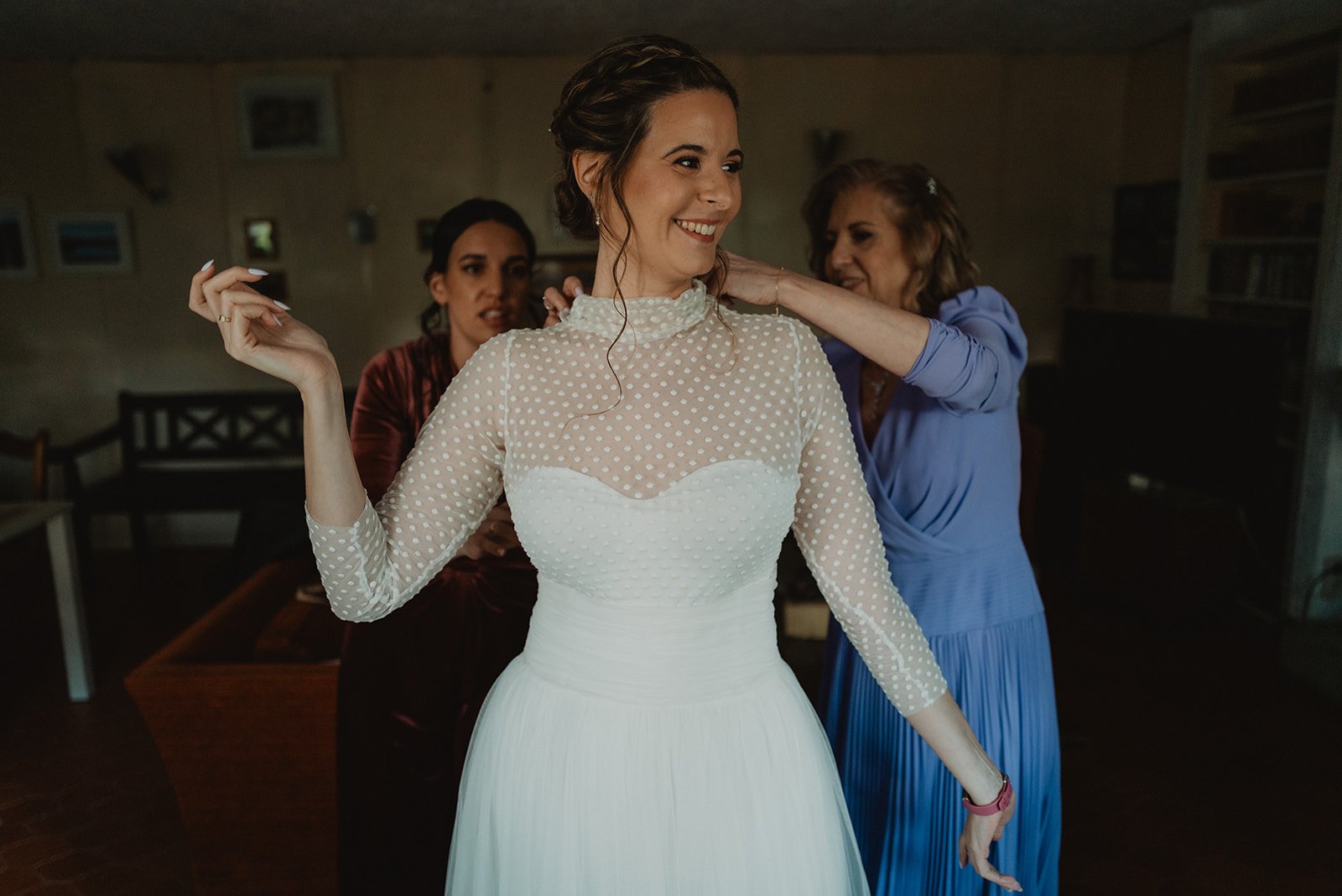 Una mujer sonriendo con vestido de novia blanco, rodeada de otras dos mujeres ayudándole a arreglarse, en un ambiente interior.