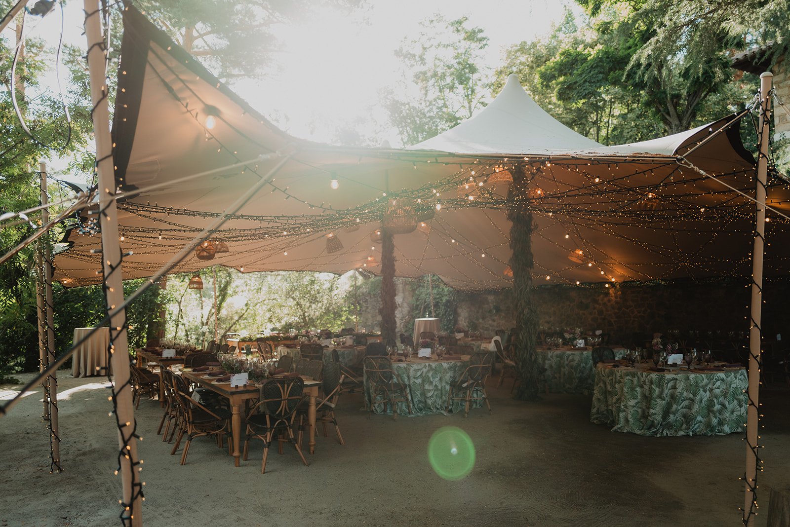 Carpa al aire libre decorada con luces para un evento, con mesas y sillas dispuestas debajo de ella, rodeado de vegetación.
