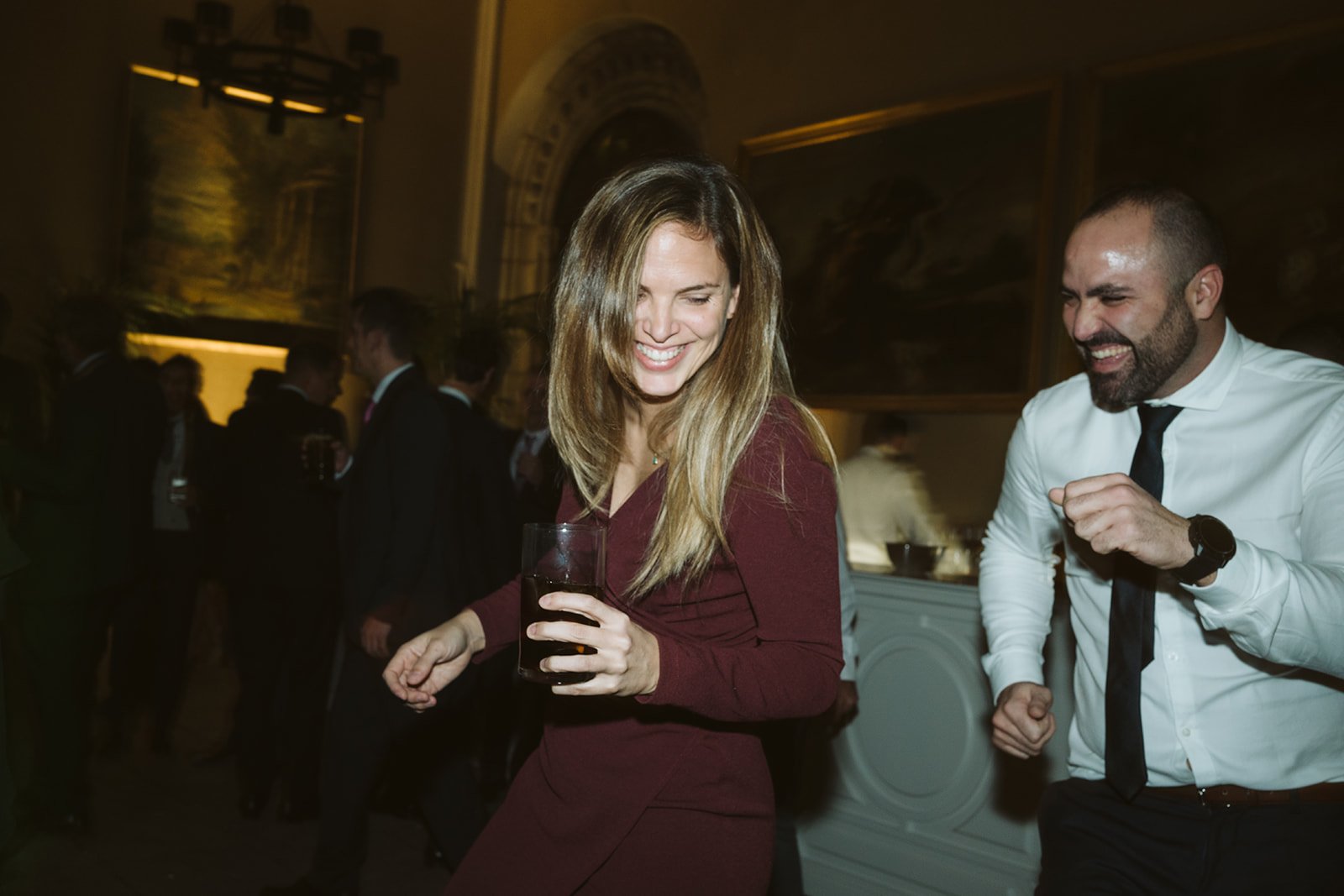 Personas bailando y riendo en una fiesta elegante, con bebidas en la mano. Ambiente festivo y formal.