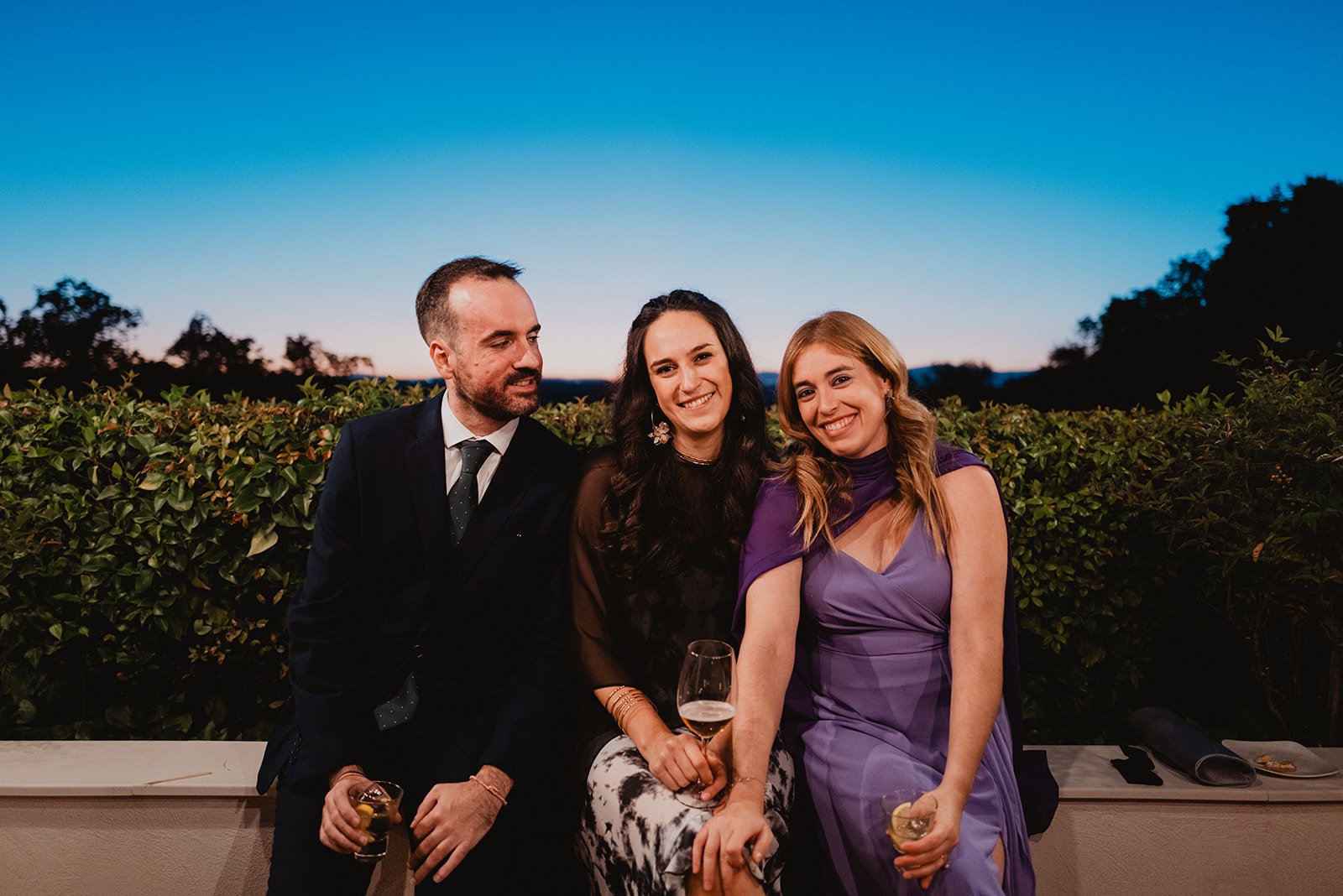 Tres personas sentadas al aire libre en una terraza, al atardecer. La persona a la izquierda lleva un traje oscuro, mientras las otras dos personas visten ropa elegante de colores oscuros y lilas. Están sonriendo y sostienen bebidas.