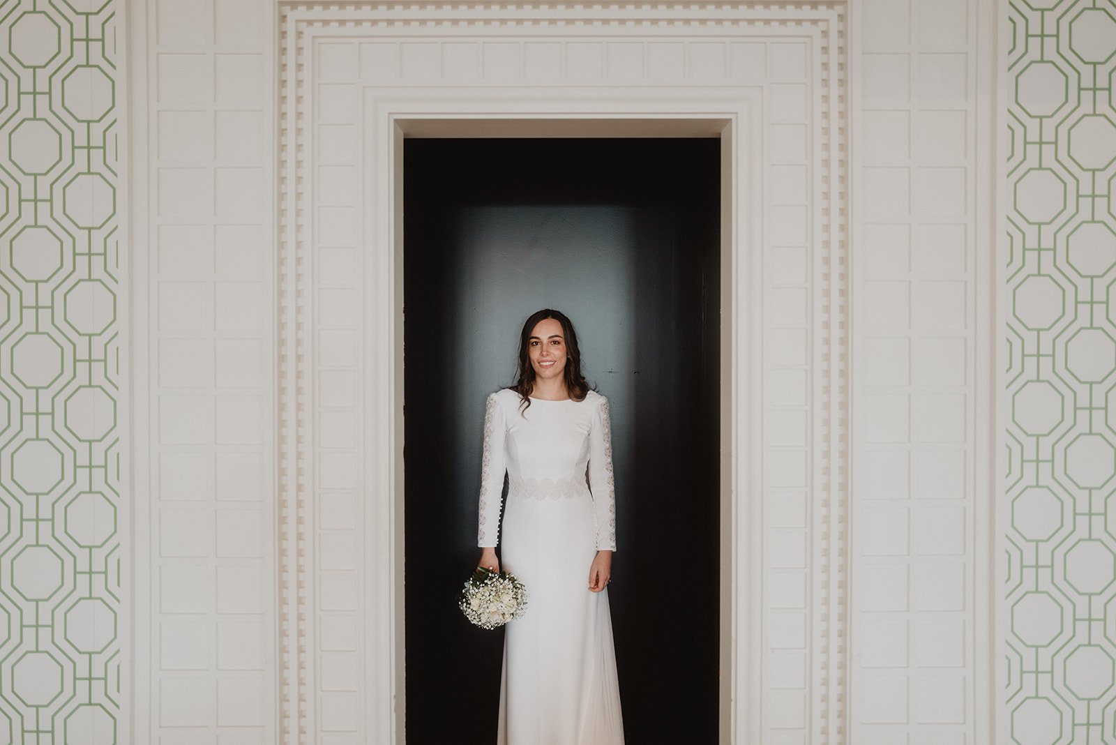 Novia en vestido blanco sosteniendo un ramo, de pie en una puerta, con fondo de papel tapiz geométrico.